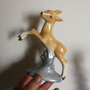 May include: A ceramic figurine of a light brown deer standing on its hind legs on a blue and white base. The deer has black eyes and a gold accent on its hooves.