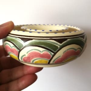 May include: A small, cream-colored ceramic bowl with a double rim. The bowl is decorated with a stylized floral pattern in shades of pink, green, yellow, and brown.