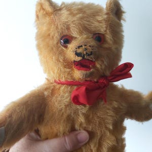 May include: A vintage teddy bear with golden fur, red eyes, and a red bow tie. The bear has a stitched nose and mouth, and its arms are outstretched. A hand is holding the bear.