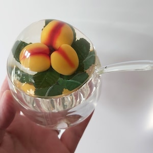 May include: A clear glass jar with a lid featuring three yellow and red peach-shaped decorations nestled among green leaves. The jar has a matching clear glass handle extending from the side, creating a cohesive design for this kitchen container.