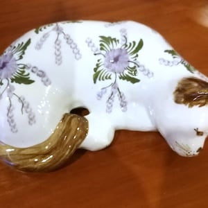May include: A ceramic cat figurine in a resting pose. The white body is decorated with purple floral designs and green leaves. The ears and tail are brown. The cat is lying down on a brown surface.