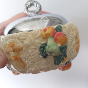 Peut inclure: Un plat de service en céramique avec un couvercle et une poignée argentés. Le plat est de couleur crème avec un motif de panier tressé et est décoré de fruits colorés. Le couvercle est brillant et réfléchissant.