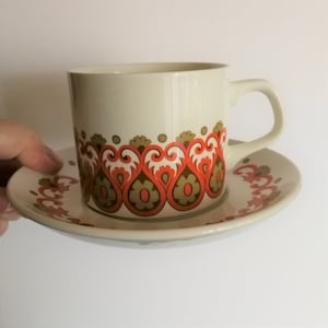 May include: Cream-colored teacup and saucer set with a repeating floral design in orange, brown, and gold. The cup has a handle and the saucer is round. A hand is holding the set.