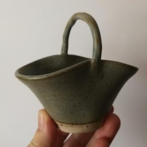 Può includere: Un piccolo cestino in ceramica fatto a mano con un manico curvo. Il cestino ha un bordo svasato e smerlato e una smaltatura verde-grigia tenue. La base è in ceramica più chiara, non smaltata. Il manico si incurva sopra l'apertura.