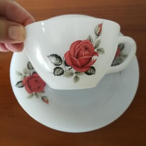 May include: White teacup and saucer set with a red rose floral design. The teacup has a handle and is placed on top of the saucer. The rose design includes red roses, green leaves, and rose buds. The set is likely made of ceramic.