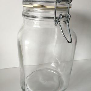 Op de afbeelding: Een heldere glazen pot met een vierkante basis en een scharnierend deksel, vastgezet met een metalen sluiting. De pot is leeg en schoon. Geschikt voor het bewaren van droge goederen. Hoogte ongeveer 15 cm.