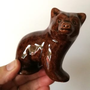 Peut inclure: Figurine d'ours en céramique marron avec une finition brillante. L'ours est en position assise, les pattes avant légèrement relevées. La figurine présente des détails, notamment un petit nez et des oreilles. Objet décoratif.
