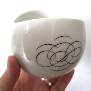 Könnte beinhalten: Eine weiße Keramikschale mit gebogenem Griff und schwarzem, abstraktem Design. Die Schale wird in einer Hand gehalten und wirkt glatt und glänzend. Das Design besteht aus Schleifenlinien.
