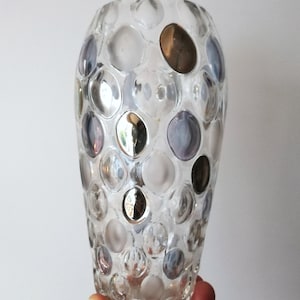 May include: Clear glass vase with a pattern of raised, irregularly sized circles in silver, white, and brown. The vase is cylindrical with a slightly flared top and is held by a hand.