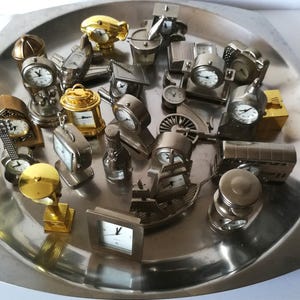 Puede incluir: Una colección de relojes en miniatura de varias formas, incluyendo una guitarra, un barco y un tren, dispuestos sobre una bandeja plateada. Los relojes son de metal y tienen esferas blancas con números y manecillas negras. Algunos tienen detalles dorados.
