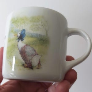 Może przedstawiać: Biały ceramiczny kubek z akwarelową ilustracją kaczki w niebieskim berecie. Kaczka jest przedstawiona na polu w zielonych i żółtych odcieniach. Kubek ma zakrzywiony uchwyt i gładkie, błyszczące wykończenie.