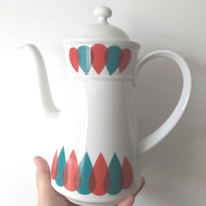 May include: White ceramic coffee pot with a lid and handle. The pot is decorated with a pattern of overlapping red and teal teardrop shapes. The pot is held by a hand.