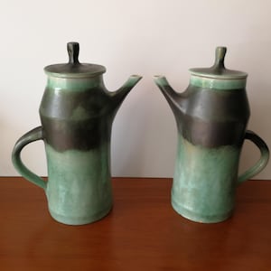 May include: Two ceramic teapots with lids. The teapots are green and black with a handle and spout. The teapots are on a wooden surface.