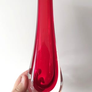 May include: A tall, teardrop-shaped glass vase featuring a vibrant, deep red centre encased in a thick layer of clear glass. The vase has a narrow, cylindrical neck that widens into a rounded, bulbous base, creating a smooth, polished appearance.