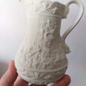May include: A small, off-white porcelain pitcher with a detailed relief design. The pitcher features a curved handle, a spout, and intricate patterns depicting classical scenes. The overall design is ornate and elegant, suitable for decorative use.