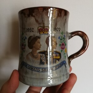 May include: A brown and white ceramic mug with a silver jubilee design. The mug features a portrait of Queen Elizabeth II and Prince Philip, along with the dates 1952 and 1977. The text "The Queen's Silver Jubilee" is printed on the mug.