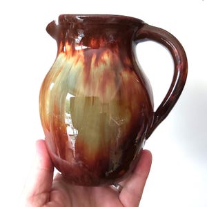 May include: A handcrafted ceramic pitcher with a glossy finish. The pitcher features a dark brown rim and handle, transitioning to a blend of red, green, and brown hues on the body. The pitcher has a spout for pouring.