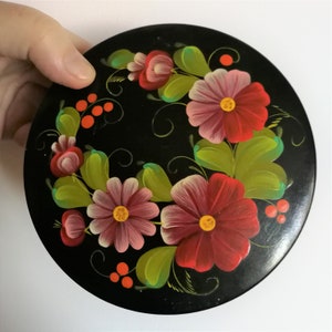 May include: A round wooden box with a black background and a hand-painted floral design. The flowers are red, pink, and white with green leaves and black swirls. There are also small red berries painted on the box.