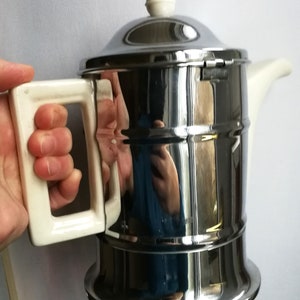 Könnte beinhalten: Eine silberne und weiße Teekanne mit Deckel und Henkel. Die Teekanne hat eine glänzende, metallische Oberfläche und einen weißen Keramikgriff.