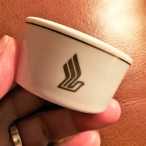 May include: A white ceramic ashtray with a gold trim and a black logo. The ashtray is held in a hand and has a curved shape.