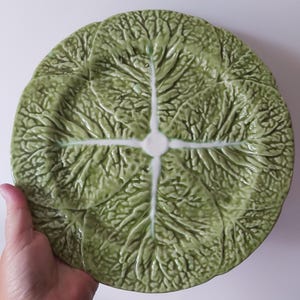 Może przedstawiać: Okrągły ceramiczny talerz o teksturowanej, tłoczonej powierzchni, która naśladuje wygląd nakładających się na siebie zielonych liści kapusty. Wzór zawiera centralny biały układ żyłek, które rozchodzą się promieniście na zewnątrz, tworząc realistyczny botaniczny wygląd.