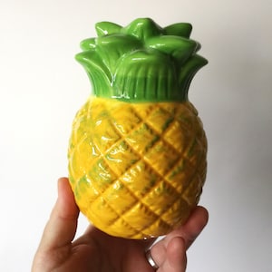 May include: A ceramic pineapple ornament with a bright yellow body and green leafy top. The pineapple has a textured surface and is held in a hand against a white background. The ornament is a decorative item.