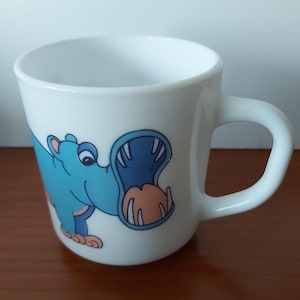 May include: White ceramic mug featuring a cartoon hippo with a wide open mouth. The hippo is blue with a peach-colored mouth and is set against a white background. The mug has a curved handle.