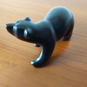 Pode incluir: Uma estatueta de urso preto, lisa e sólida, representada em posição de pé. A escultura apresenta uma forma arredondada e estilizada com um acabamento brilhante, orelhas e focinho bem definidos, capturando o animal numa postura frontal.