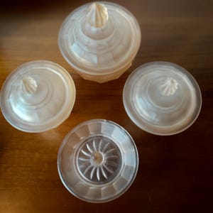 May include: Four clear, decorative glass containers with lids. The lids have a textured, conical finial. The containers are arranged on a wooden surface. The containers have a faceted design.