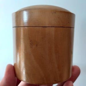 May include: A cylindrical wooden box with a domed lid. The box is made of light brown wood and has a smooth, polished finish. The lid fits snugly on top of the box. The box is held in a person's hand.