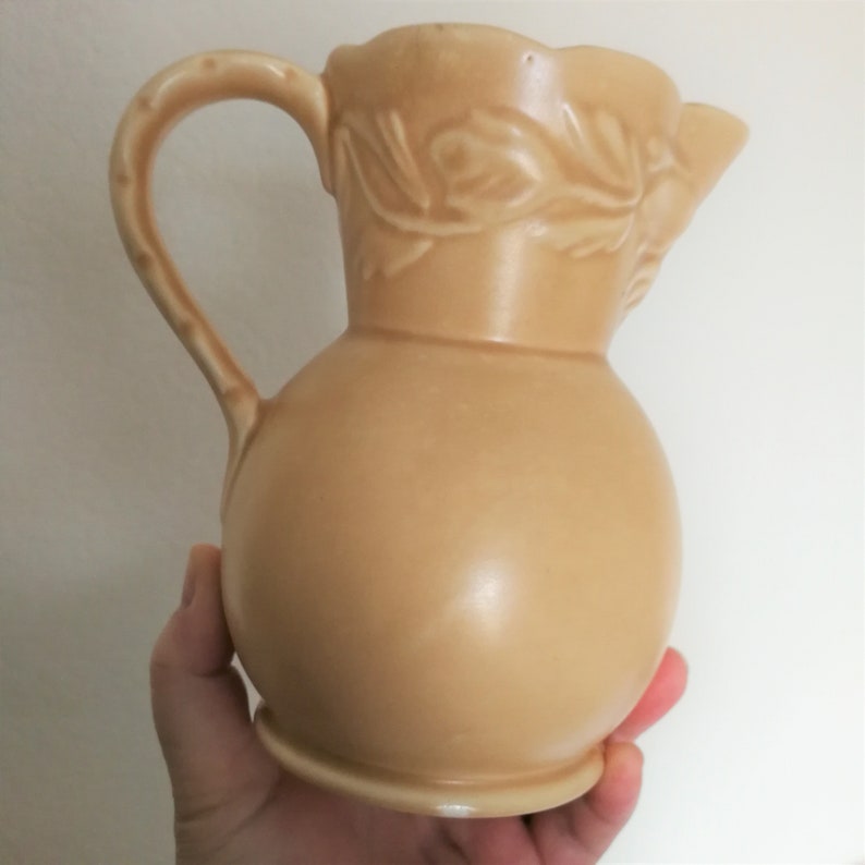 May include: A beige ceramic pitcher with a floral design. The pitcher has a round body and a curved handle.