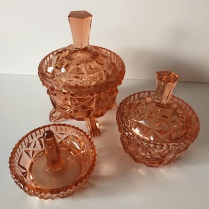 Könnte beinhalten: Ein Set aus drei vintage, pfirsichfarbenen Glasbehältern. Das Set beinhaltet ein Gefäß mit Deckel und geometrischem Muster, ein kleineres Gefäß mit Deckel und einen Ringhalter mit zentralem Pfosten.