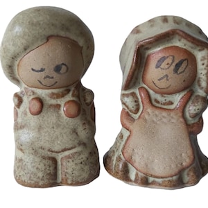 Può includere: Due statuine in ceramica, un ragazzo e una ragazza, con finitura rustica e macchiettata. Il ragazzo ha la testa rotonda e un occhio che ammicca. La ragazza indossa un cappello e un grembiule. Entrambi hanno volti sorridenti e sono di colore marrone chiaro.