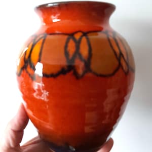 May include: A ceramic vase with a glossy, orange glaze. The vase has a decorative pattern in shades of orange and black. The vase is held in a hand.