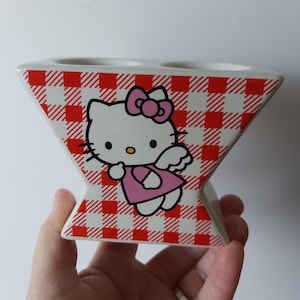 May include: A white ceramic toothbrush holder with a red and white checkered pattern. The holder features a Hello Kitty graphic in a pink dress and bow. The holder has two openings for toothbrushes.
