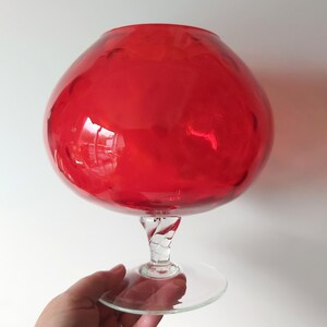 May include: A large, vibrant red glass goblet with a rounded bowl and a clear, twisted stem. The goblet sits on a clear, circular base. The glass reflects light, creating highlights and shadows.