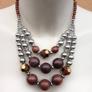 May include: A multi-strand necklace featuring brown wooden beads, silver beads, and gold-toned beads. The necklace is layered and has a bohemian style.