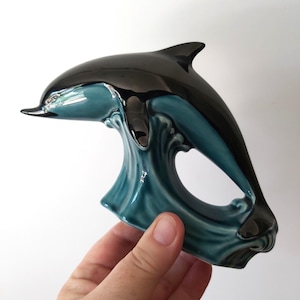 May include: A glossy ceramic figurine of a dolphin leaping from a stylized wave. The dolphin is finished in a deep black glaze, while the wave base features a contrasting teal blue glaze with a smooth, reflective surface and curved details.