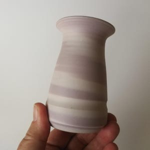 Può includere: Un vaso in ceramica fatto a mano con un design a strati unico. Il vaso presenta fasce alternate di viola chiaro e bianco sporco, creando un motivo a righe. Il vaso ha una parte superiore svasata e un corpo leggermente affusolato.