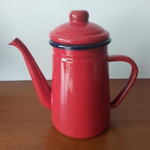 May include: A bright red enamel coffee pot with a matching lid and a dark blue rim. The pot features a long, curved spout on the left side and a sturdy handle on the right side. The surface is smooth and glossy, showcasing a classic cylindrical shape.