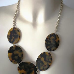 Puede incluir: Collar con grandes colgantes ovalados de carey, unidos por una cadena dorada. Los colgantes muestran un patrón moteado marrón y amarillo, creando un accesorio audaz y elegante. Diseño clásico.