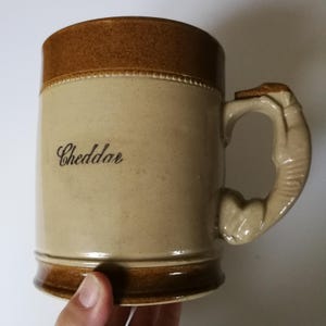 May include: A brown and beige ceramic beer stein with a handle shaped like a dog's paw. The stein has the word "Cheddar" written on the front.