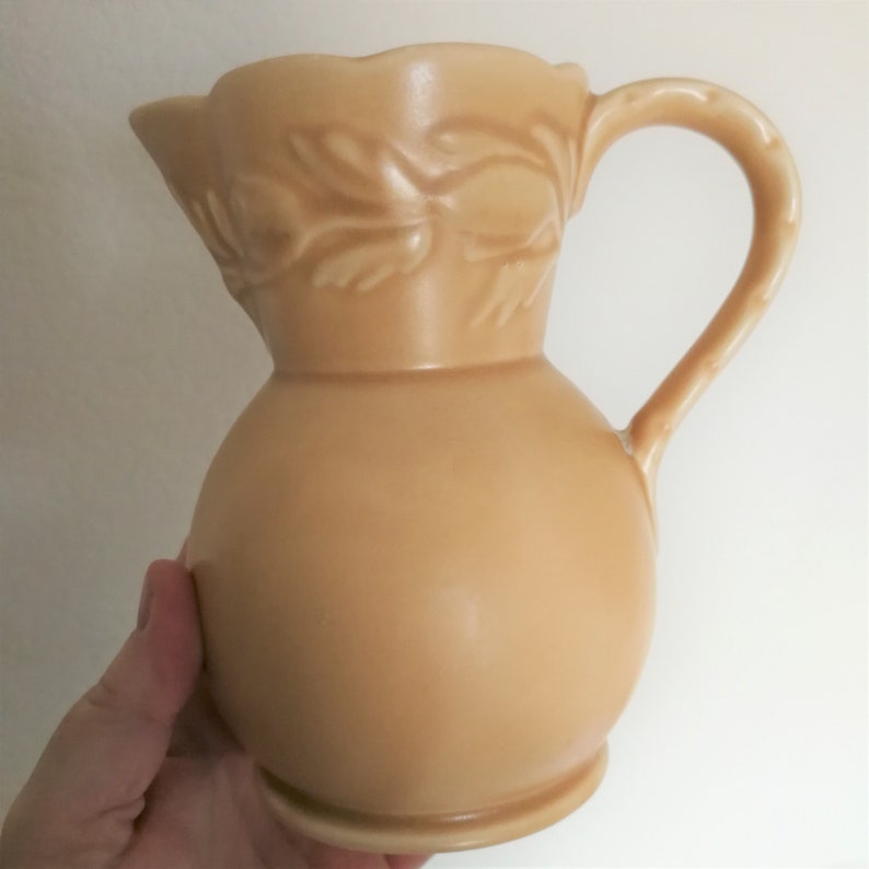 May include: A beige ceramic pitcher with a floral design. The pitcher has a round body and a curved handle.