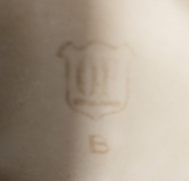 May include: Close-up of a white porcelain mark with a shield-shaped logo containing the letters "GF" and the letter "B" below it.