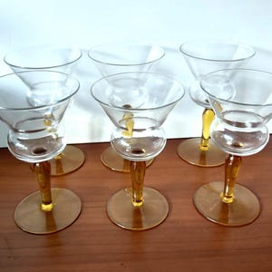Puede incluir: Seis copas de martini transparentes con tallos y bases de color ámbar. Las copas tienen una forma de cuenco cónico clásico y un diseño de tallo de doble burbuja. Las bases son redondas y los tallos son delgados.
