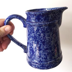 Op de afbeelding: Een blauw-wit gespikkelde keramische kan met een handvat en tuit. De kan heeft een klassiek ontwerp met een afgeronde body en een stevig handvat. De blauwe kleur is een diepe tint en de witte spikkels voegen een unieke textuur toe.