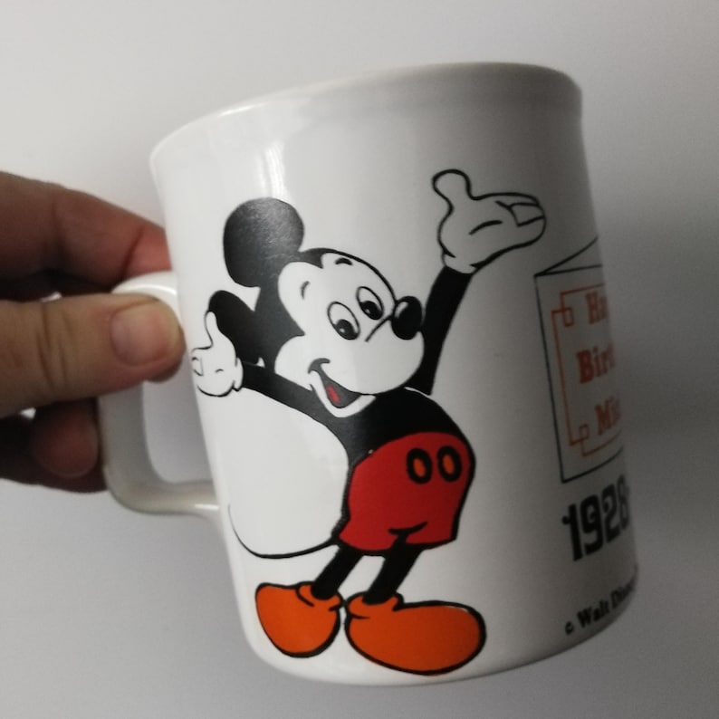 K&ouml;nnte beinhalten: Wei&szlig;er Keramikbecher mit einer Abbildung von Mickey Mouse in Schwarz, Rot und Orange. Der Becher hat einen Henkel und tr&auml;gt den Text "Happy Birthday Mickey" und das Jahr "1928".