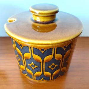 May include: A ceramic sugar bowl with a glossy brown glaze and a matching lid. The exterior features a repeating dark brown geometric pattern of stylised arches and diamond shapes. A small notch is cut into the rim to accommodate a spoon.