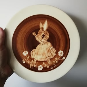 Może przedstawiać: Zdobiony talerz w stylu vintage w brązowo-białej kolorystyce. Środek talerza przedstawia uroczą ilustrację królika w sukience, otoczonego kwiatowymi akcentami. Talerz ma białą obwódkę.