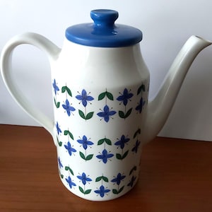 May include: White ceramic coffee pot with a blue lid and handle. The pot is decorated with a repeating pattern of blue flowers and green leaves. The spout is long and curved.